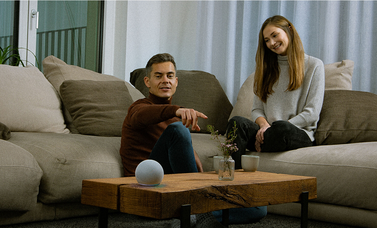 A man and a woman, a seemingly happy couple, are sitting comfortably on their couch in what presumably is their living room. The man is pointing towards something while looking at a Voice Assistent on their couch table, having given it a task probably.