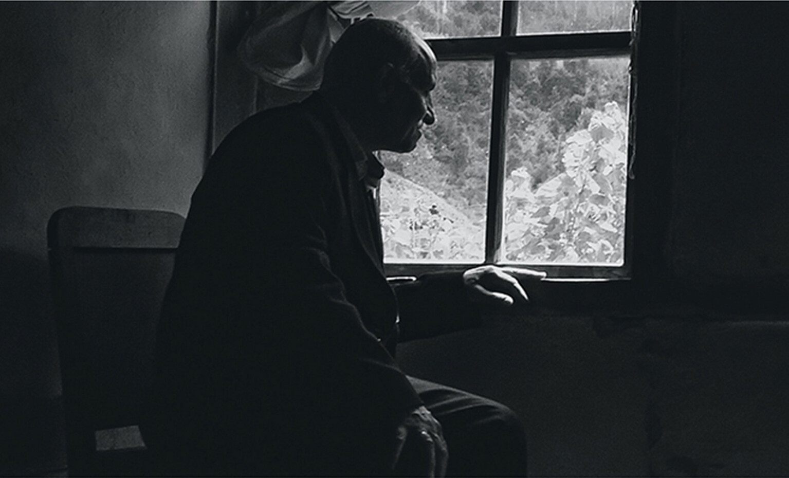Black and white photograph of an elderly man in a dark corner of a house, looking out of the window. His hand is resting on the windowsill, almost like wanting to get in touch with the world outside.