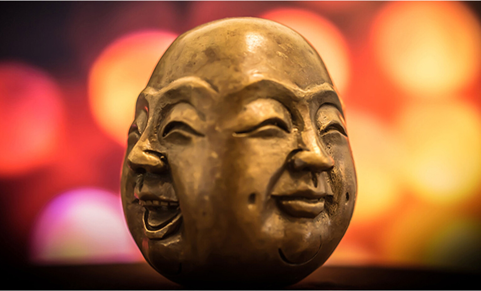 Leuchtende und bunte Fotografie einer Buddha Statue. Die Statue ist fast schon Ei-förmig und stellt die vier Gesichter Buddhas rundherum dar (lächelnd, lachend, weinend, wütend).