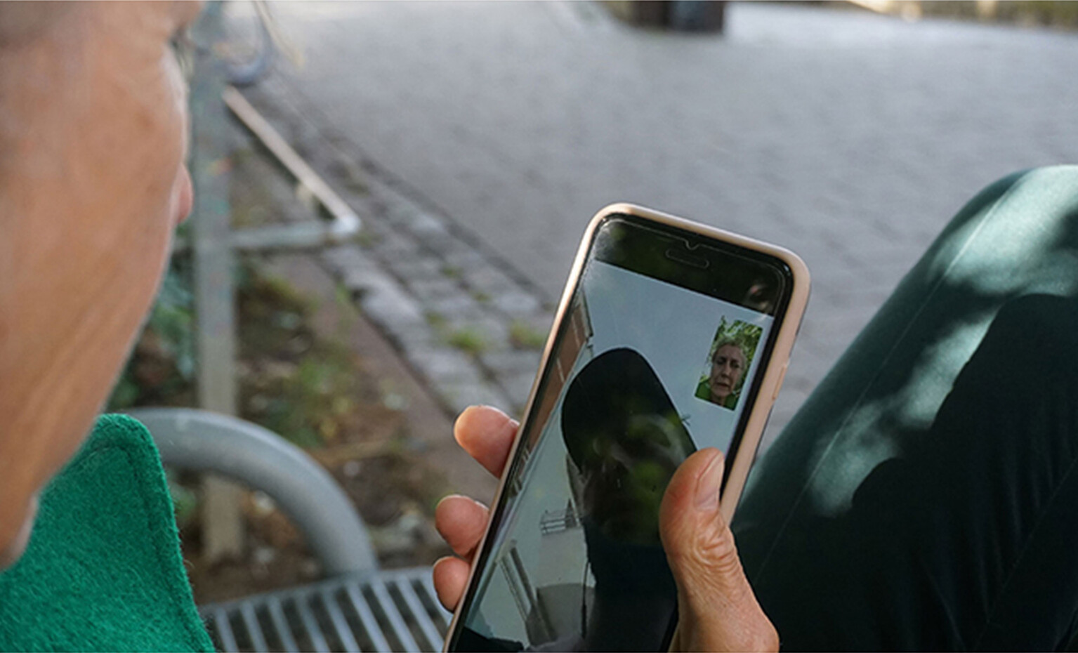 Elderly woman sitting on a bench in what seems to be a park with a mobile phone in her hand. She is on a videocall with a younger person..