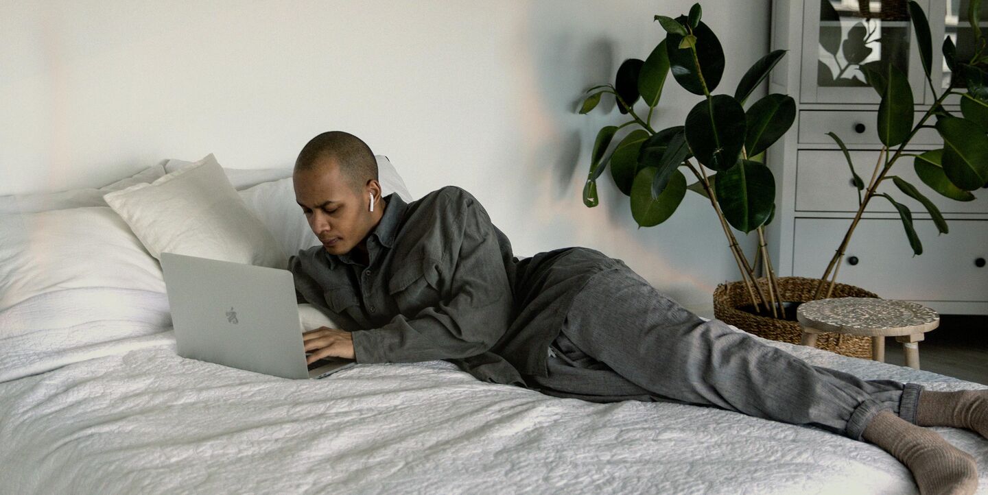 VUI.agency Finance Industries – Conversational Banking – dark skinned bold man in grey clothes lying on his bed with white sheets  using a macbook
